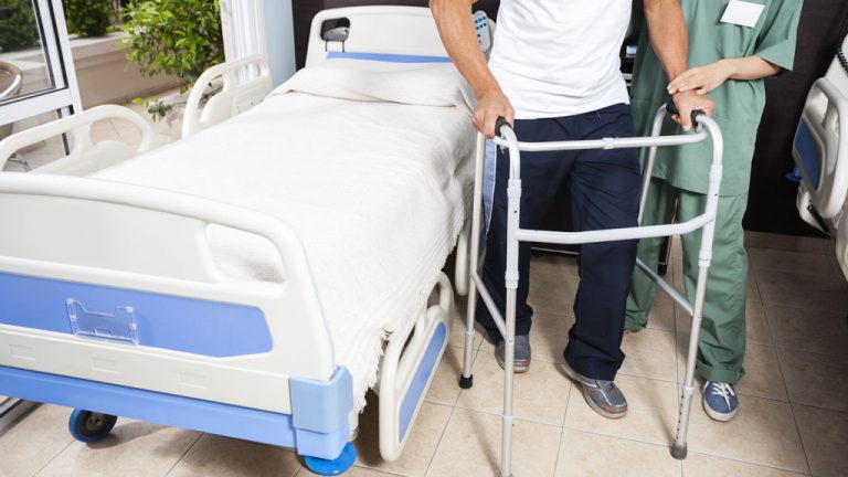 A man using a walker stands next to a hospital bed and is being helped by a nurse.