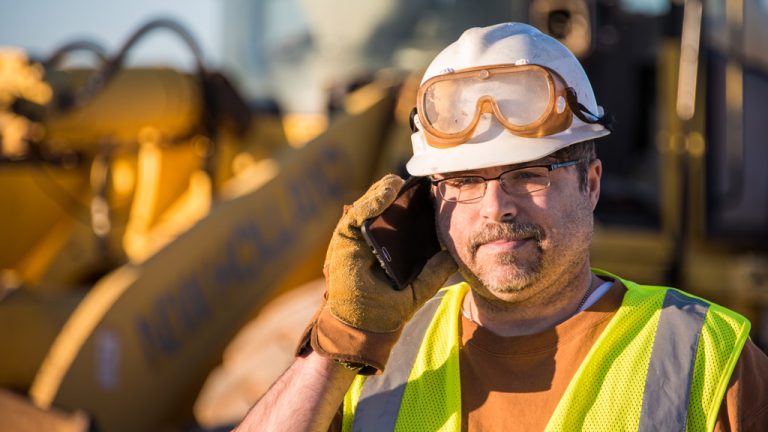 A construction worker stands in front of a large machine while talking on the phone.