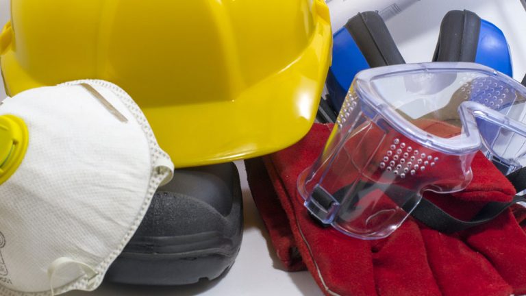 A photo of various construction safety equipment. It includes hearing protection, safety googles, gloves, a hardhat, boots and a surgical mask.