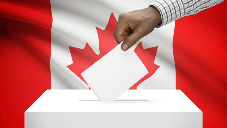 A hand places a slip of paper into the slit of a white box with the Canadian flag in the background.