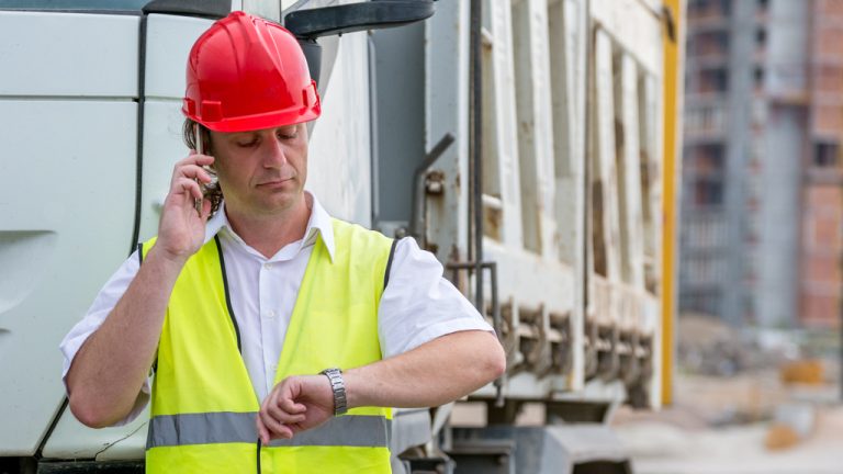A construction worker standing in front of a truck checks his watch while talking on the phone.