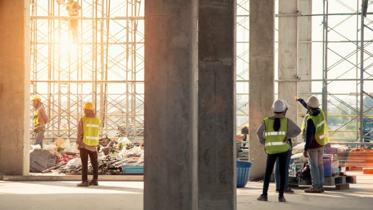 Stone pillars in the foreground divide the image in half with construction workers on either side on a construction site.