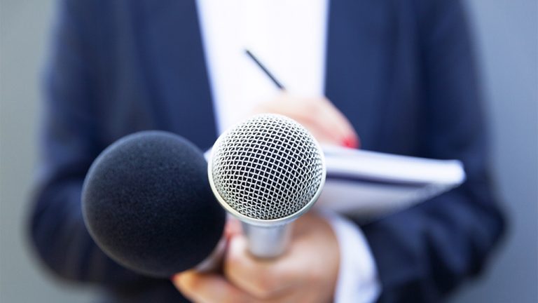 a reporter writing in a notebook while holding out two microphones.