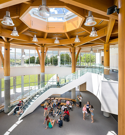 The Southern Okanagan Secondary School double-height mass timber atrium showcases biophilic design principles, with its abundant natural light, views to nature and tree-like exposed concrete-wood columns supporting a hexagonal arrangement of visible glulam roof  beams.